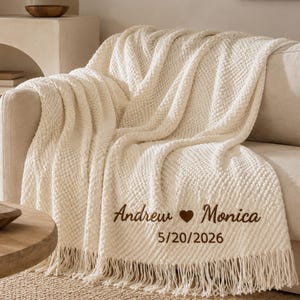 Puede incluir: Una manta texturizada de color crema con flecos, que presenta el texto bordado en marrón Andrew corazón Monica y la fecha 20/05/2026. La manta tiene un patrón de puntos en relieve repetido en toda su superficie y está sobre un sofá.