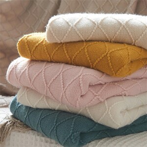 May include: A stack of five folded blankets in various colors: cream, mustard yellow, light pink, off-white, and teal. Each blanket features a diamond pattern. The blankets are soft and cozy, perfect for adding warmth and style to any space.