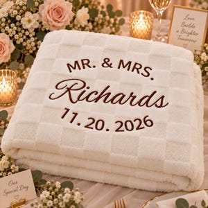 Pode incluir: Uma manta branca com textura xadrez e bordado em castanho escuro que diz MR. & MRS. Richards e a data 11.20.2026. A manta tem um aspeto macio e felpudo com um padrão de quadrados repetitivos em toda a superfície.