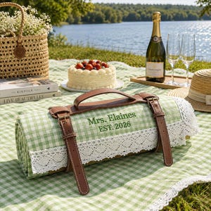 May include: A rolled picnic blanket featuring a light green and white gingham pattern, trimmed with white lace, and secured by brown leather straps with a handle. The fabric is embroidered with the green text: Mrs. Elaines EST. 2026.