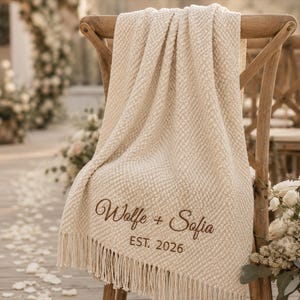 May include: A cream-colored textured throw blanket with fringe trim draped over a wooden chair. The blanket features brown embroidered text that reads "Wolfe + Sofia" above the text "EST. 2026" in a clean, sans-serif font style.