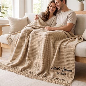 Può includere: Una coperta in maglia testurizzata color beige con bordi sfrangiati. La coperta presenta un testo ricamato in nero che recita Mark + Jeanne est. 2026. Il tessuto ha un motivo a punti in rilievo che si ripete su tutta la superficie.