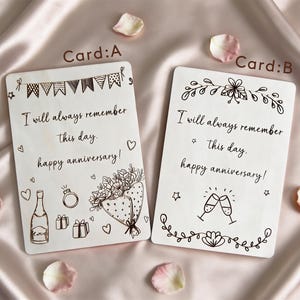May include: Two white anniversary cards with black ink illustrations and text. Card A has a banner, champagne bottle, ring, gifts, and a bouquet. Card B has floral designs and champagne glasses. Both cards read "I will always remember this day. Happy anniversary!"