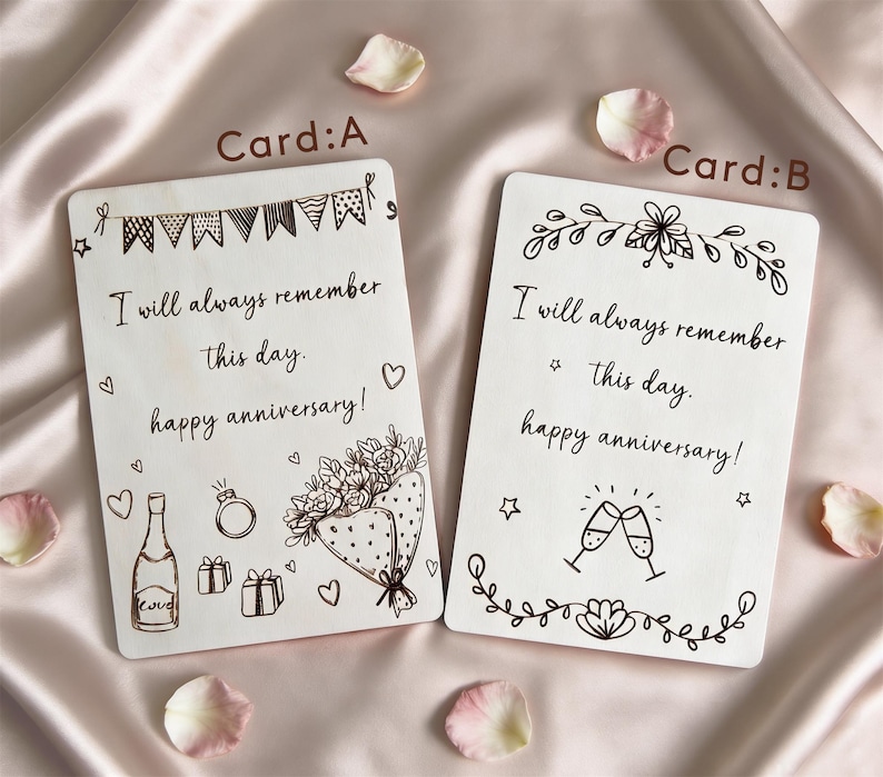 May include: Two white anniversary cards with black ink illustrations and text. Card A has a banner, champagne bottle, ring, gifts, and a bouquet. Card B has floral designs and champagne glasses. Both cards read "I will always remember this day. Happy anniversary!"