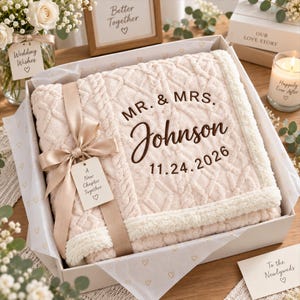 Op de afbeelding: Een crèmekleurige deken met kabelstructuur, gevouwen in een witte geschenkdoos, vastgebonden met een beige satijnen lint en een label met de tekst A New Chapter Together. De deken heeft donkerbruin borduurwerk met MR. & MRS. Johnson 11.24.2026.