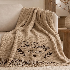 May include: A beige knit throw blanket featuring a textured pattern and fringe trim. The blanket is embroidered with the dark brown text "The Timothys", "EST. 2026", a small heart icon, and two leafy branch motifs centered on the lower portion.