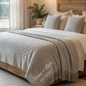 May include: A light gray throw blanket with a diamond pattern and fringe detail is draped over a wooden bed frame. The blanket is embroidered with the names "James + Beth" and the year "EST 2026". Two matching pillows are on the bed.