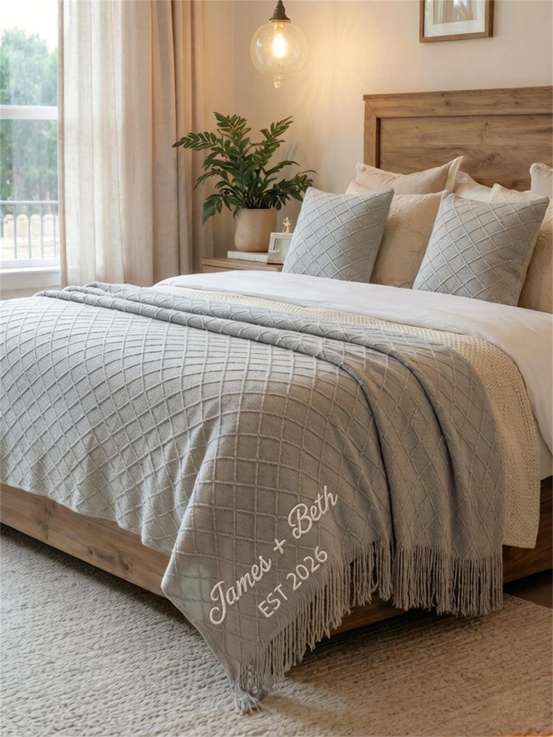 May include: A light gray throw blanket with a diamond pattern and fringe detail is draped over a wooden bed frame. The blanket is embroidered with the names "James + Beth" and the year "EST 2026". Two matching pillows are on the bed.