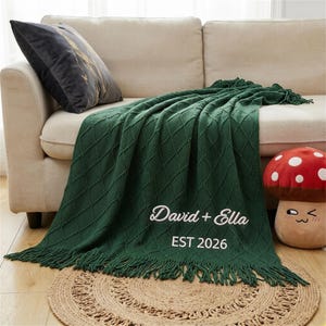 May include: A dark green knitted throw blanket with a diamond pattern and fringe, draped over a beige sofa. The blanket has the embroidered names "David + Ella" and the year "EST 2026". A decorative pillow and mushroom-shaped plush toy are also visible.