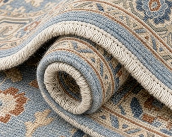 May include: A rolled area rug featuring a light blue base with intricate floral and geometric patterns in shades of beige, tan, and dark blue. The rug has a thick, woven texture with a finished edge and a light beige fringe along the border.