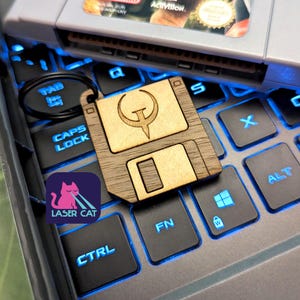 May include: A wooden floppy disk keychain with the Quake logo, resting on a backlit keyboard. A Super Nintendo game cartridge is in the background. The Laser Cat logo is visible on the keyboard.