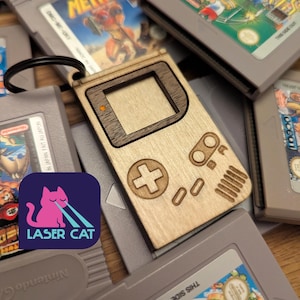 May include: A wooden keychain shaped like a classic handheld video game console, featuring engraved buttons and a screen. The keychain is light-colored with a darker wood accent. Surrounding the keychain are several gray game cartridges. A pink cat logo with the text "LASER CAT" is in the lower left.