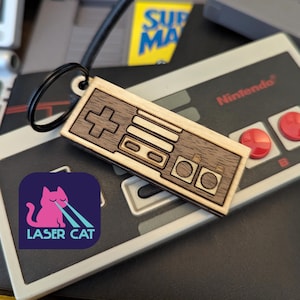 May include: A wooden keychain in the shape of a retro games controller, with a cross-shaped D-pad and rectangular buttons. The keychain is attached to a black ring. The Laser Cat logo is visible in the lower left corner.