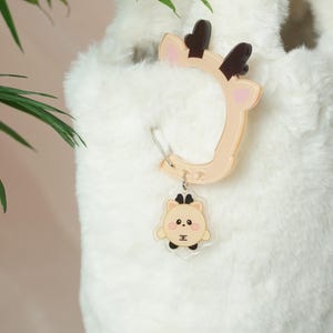 May include: A beige and brown deer-shaped keychain and a matching deer-shaped ring. The keychain features a cartoon deer face with a crown. The ring is in the shape of a deer head with antlers. The items are attached to a fluffy white surface.