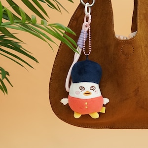 May include: A plush penguin keychain with a navy blue hat, red shirt, and yellow feet. The keychain is attached to a brown suede bag with a silver metal ring. The penguin has embroidered eyes and a small yellow tag.