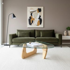 Puede incluir: Un salón moderno con un sofá de terciopelo verde y cojines a juego. Una mesa de centro con tapa de cristal y base de madera descansa sobre una alfombra blanca. Una pintura abstracta cuelga sobre el sofá y una lámpara de pie negra está en la esquina.