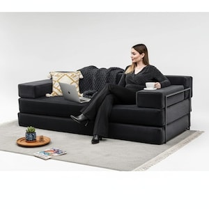 Könnte beinhalten: Ein schwarzes Modulsofa mit Metallrahmen und integrierter Armlehne. Eine Frau sitzt auf dem Sofa und hält eine Tasse. Ein Laptop, ein dekoratives Kissen und eine Decke befinden sich auf dem Sofa. Ein kleiner Teppich und ein Holztablett mit einer Pflanze sind davor.