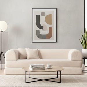 Pode incluir: Uma sala de estar moderna com um sofá creme e almofadas a condizer. Uma mesa de centro redonda de madeira com base de metal preta suporta livros e uma chávena de chá. Uma impressão de arte abstrata está pendurada acima do sofá. Uma luminária de chão e uma planta em vaso também são visíveis.