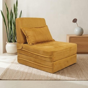 May include: Mustard yellow corduroy convertible chair with a matching cushion. The chair is folded and has a ribbed texture. A potted plant and a small wooden cabinet are in the background. The chair is designed for lounging.