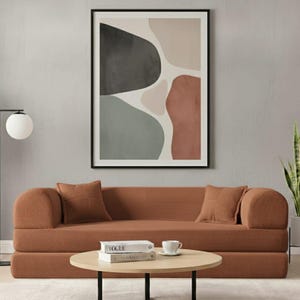 May include: A contemporary living room scene with a terracotta-coloured sofa and two square cushions. A round wooden coffee table with a black metal frame holds books and a teacup. An abstract art print hangs above the sofa.