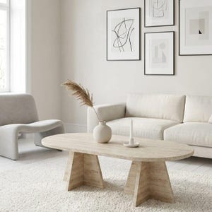 Op de afbeelding: Een ovale salontafel van beige travertijn met een getextureerd stenen oppervlak en twee brede, schuine voetstukken. De tafel staat op een wit tapijt en bevat een kleine ronde keramische vaas met een gedroogde plantenstengel en een witte kaars.