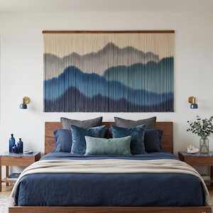 May include: A bedroom with a wooden bed frame, navy blue bedding, and decorative pillows. A large wall hanging depicts a mountain range in shades of blue and gray. Two nightstands with blue lamps and decor flank the bed.