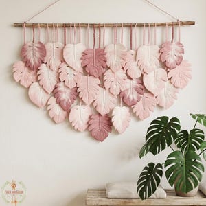 May include: A decorative macrame wall hanging with a collection of pink and white leaf-shaped elements. The leaves are suspended from a wooden dowel, creating a textured, cascading design. The wall hanging is displayed against a white wall, adding a touch of bohemian style.
