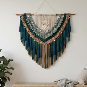 May include: A large, handcrafted macrame wall hanging in teal, green, and tan. The textile art piece features a wooden dowel, intricate knotting, and cascading tassels. The design includes a central geometric pattern and beaded accents, creating a bohemian aesthetic.