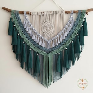 Peut inclure: Suspension murale en macramé faite à la main, dans des tons de vert, de bleu et de crème, suspendue à une branche en bois naturel. Le design présente des nœuds complexes, des glands et des perles, créant une pièce décorative d'inspiration bohème.