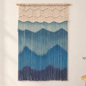 May include: A wall hanging featuring a wooden dowel at the top with cream-colored macrame cord arranged in wavy patterns. Below the macrame, long vertical threads transition in color from light blue to deep indigo in a mountain-like design.