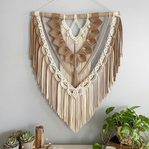 以下が含まれることがあります： クリーム、ベージュ、茶色の色合いの大きなマクラメの壁掛け。手作りのテキスタイルは、複雑な結び目のパターンとフリンジが特徴で、天然の木のダボから吊り下げられています。デザインは対称的で幾何学的で、ボヘミアンな美学を作り出しています。