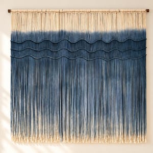 May include: A large macrame wall hanging featuring a cream-colored top section that transitions into a deep indigo blue ombre gradient. Three horizontal, wavy dark blue ropes are woven across the center of the long, vertical fringe.