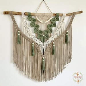 Könnte beinhalten: Ein großer Makramee-Wandbehang mit einem Treibholzast. Das Design umfasst beige, weiße und olivgrüne Knotenmuster mit Fransen und Quasten. Das Stück ist eine dekorative Textilkunst, geeignet für die Inneneinrichtung.