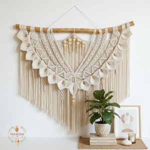 Può includere: Un grande arazzo da parete in macramè color crema con un'asta di legno e perline decorative in legno. Il macramè presenta intricati motivi di nodi e un bordo con frange. L'arazzo è esposto su una parete bianca.