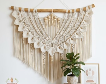 May include: A large, cream-colored macrame wall hanging with a wooden dowel and decorative wooden beads. The macrame features intricate knotting patterns and a fringed bottom. The wall hanging is displayed on a white wall.