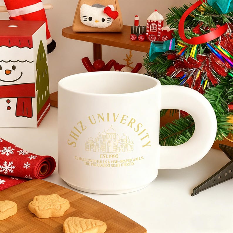 Pode incluir: Uma caneca de cer&acirc;mica branca com uma al&ccedil;a grande e circular. A caneca apresenta o texto "SHIZ UNIVERSITY" em letras douradas, juntamente com um gr&aacute;fico de um edif&iacute;cio e o texto "EST. 1995". A caneca est&aacute; em uma t&aacute;bua de corte de madeira com biscoitos.