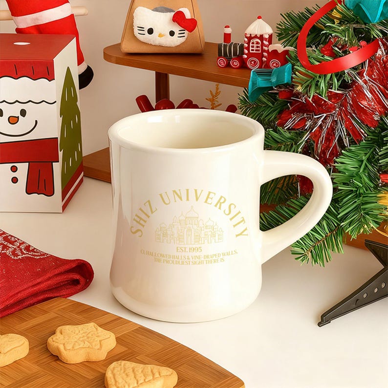 Pode incluir: Caneca de cer&acirc;mica creme com a inscri&ccedil;&atilde;o dourada "SHIZ UNIVERSITY". A caneca tem uma al&ccedil;a curva e uma base larga. A caneca est&aacute; sobre uma superf&iacute;cie de madeira com biscoitos. Decora&ccedil;&otilde;es de Natal ao fundo.