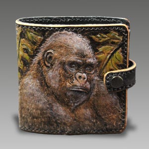 May include: A square leather wallet featuring a detailed, embossed, and hand-painted portrait of a gorilla in shades of brown and black. The wallet has a black leather border with visible stitching and a side strap secured by a dark metal snap button.