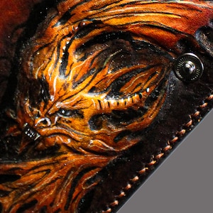 Könnte beinhalten: Nahaufnahme einer Ledergeldbörse mit einem detaillierten, handgefertigten Design einer wilden, orange-schwarzen Kreatur. Das Kunstwerk ist vor einem dunkelbraunen Hintergrund mit einem Metall-Druckknopfverschluss angeordnet.