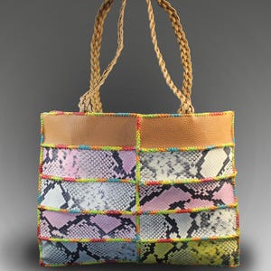May include: A colorful tote bag with a patchwork design. The bag features panels of faux snakeskin in various colors, including pink, blue, and yellow, stitched together with rainbow thread. It has a brown leather top and braided handles.