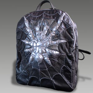 Puede incluir: Mochila de cuero negro con un diseño de telaraña plateada. La mochila tiene un asa superior, una cremallera blanca y un pequeño bolsillo lateral. El diseño de la telaraña está cosido con hilo blanco.