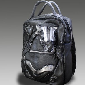 Puede incluir: Mochila de cuero negro con detalles plateados y asa superior. La bolsa presenta un diseño de patchwork con costuras visibles y un bolsillo con cremallera. La estética general es una mezcla de estilos industrial y artesanal.