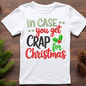 Może przedstawiać: Biała koszulka z napisem "in CASE... you get CRAP for Christmas" w zielonych, czerwonych i czarnych literach. W projekcie uwzględniono gałązkę ostrokrzewu. Koszulka jest prezentowana na drewnianej powierzchni.