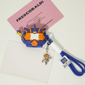 Op de afbeelding: Een blauwe en oranje beschermhoes voor draadloze oordopjes, met een robotachtig ontwerp. Het bevat een sleutelhanger met een kleine bedel en een blauw koord. De afbeelding toont ook een roze kaart met het woord "FRESCOBALDI".