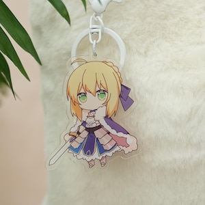 Peut inclure: Un porte-clés en acrylique transparent représentant un personnage d'anime de style chibi aux cheveux blonds et aux yeux verts. Le personnage porte une tenue bleue et blanche avec une cape violette et tient une épée argentée. Le porte-clés a un anneau en métal blanc.