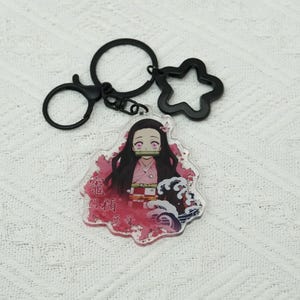 Könnte beinhalten: Ein klarer Acryl-Schlüsselanhänger mit einem Charakter aus einer Anime-Serie. Der Schlüsselanhänger hat einen schwarzen Metallring, einen sternförmigen Anhänger und einen Karabinerverschluss. Der Charakter hat lange schwarze Haare, rosa Augen und trägt einen rosa Kimono. Japanischer Text ist sichtbar.