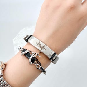 Pode incluir: Uma pulseira de metal prateado com uma placa retangular gravada com um design de espada e flores, combinada com uma segunda pulseira composta por uma pedra retangular preta numa corrente prateada com detalhes metálicos.