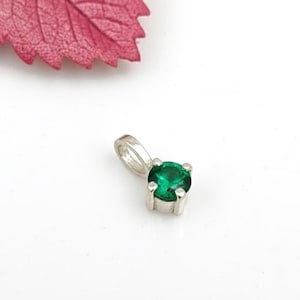 Op de afbeelding: Een kleine zilveren hanger met een ronde, smaragdgroene edelsteen. De hanger heeft een eenvoudig, elegant ontwerp, perfect voor dagelijks gebruik. De edelsteen is gezet in een vier-klauwzetting. Witte achtergrond met een rood blad.