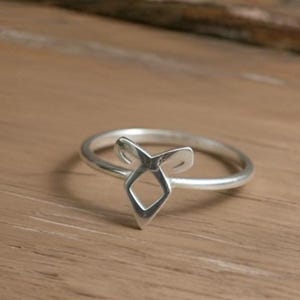 K&ouml;nnte beinhalten: Ein silberner Ring mit einem stilisierten geometrischen Design. Der Ring hat eine zentrale Diamantenform mit zwei nach oben gebogenen Elementen, die auf einem d&uuml;nnen Band angeordnet sind. Der Ring wird auf einer Holzoberfl&auml;che pr&auml;sentiert.