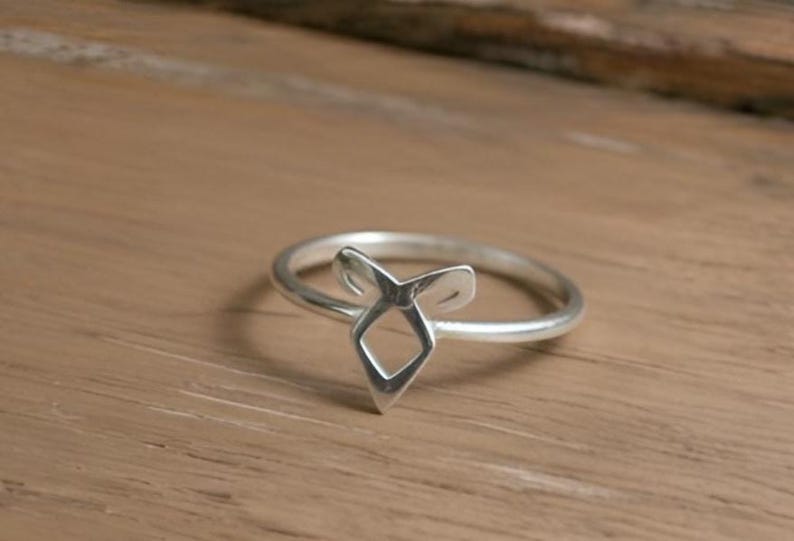 K&ouml;nnte beinhalten: Ein silberner Ring mit einem stilisierten geometrischen Design. Der Ring hat eine zentrale Diamantenform mit zwei nach oben gebogenen Elementen, die auf einem d&uuml;nnen Band angeordnet sind. Der Ring wird auf einer Holzoberfl&auml;che pr&auml;sentiert.